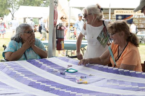 A woman in a blue shirt claps her hand over her mouth next to a purple quilt while a woman in a white blouse smiles and a woman in an orange shirt laughs.