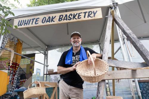 A man smiles, posing and holding a woven basket under a tent with a hand-painted sign: White Oak Basketry. 