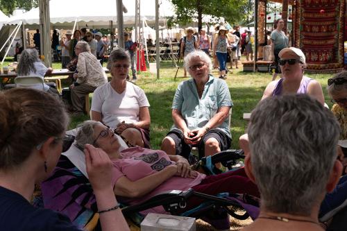 A woman lies in a reclined chair with eyes closed while people in chairs sing around her.