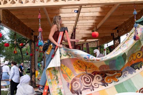 A woman on a ladder stands underneath a wooden building, holding the end of a tapestry depicting animals and trees in warm pastel colors, pointing off in the distance.