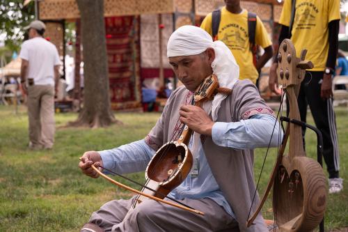  man in a headwrap and traditional Kazakh dress bows a stringed instrument with a round resonator and narrow neck, a concentrated look on his face.