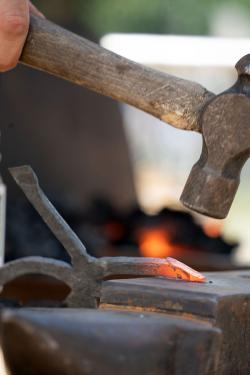 Close-up on a metal gig, one tip glowing red, against an anvil with a hammer poised above. Coals burn red in the background.