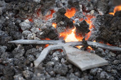 A large pile of black coal sits in a furnace. A half-formed metal gig, similar to a small trident, sits in the hottest part of the coals. The tines of the gig glow orange from the heat.