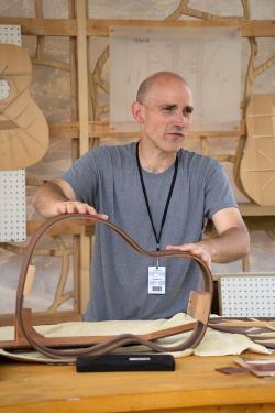A man stands behind a table and touches a piece of wood shaped like the body of a guitar. He speaks to visitors about guitar making. Behind him are more wooden pieces demonstrating the different steps of the guitar making process.