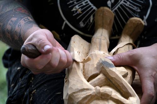  Two hands hold the body of a carved wood figure. One hand uses a tool to carve off a layer of wood while the other holds the block steady.