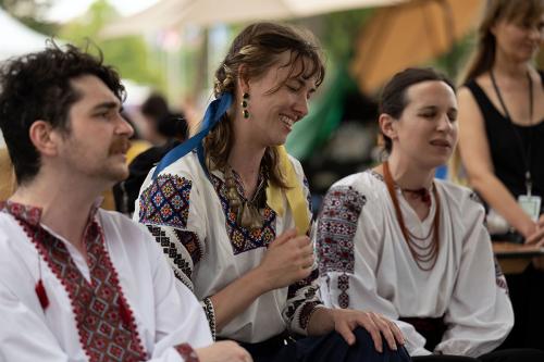 Three people, seated, sing. All wear embroidered white shirts. The woman at center, with blue and yellow ribbons in her hair, smiles widely.