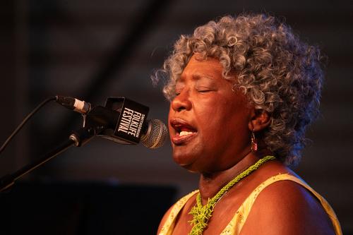 Close-up on a woman singing on stage, wearing a yellow strapless dress and green beaded necklace.