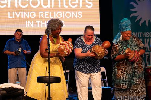 Three women play shaker gourd instruments on stage. A man plays percussion in the background.