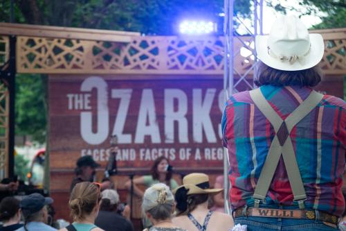 Out of focus, a couple performs on a small outdoor stage for a crowd of people. In the foreground, the back of a person wearing a cowboy hat, suspenders, and a leath belt engraved with WILLI.