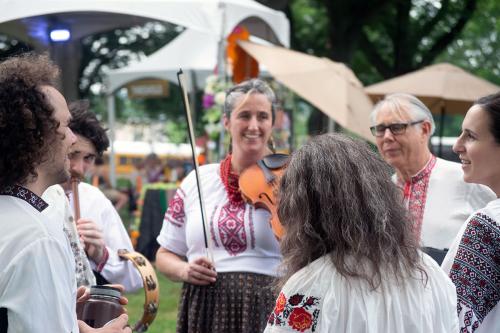 Six people stand in a circle, singing and playing instruments: violin, tamborine, and flute.