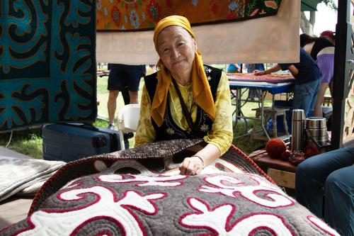 Seated outdoors, a woman smiles at the camera, a large gray wool textile with white and red appliqué work in her lap.