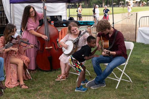 Three women and one man sit in folding chairs in a half circle on grass. They are playing instruments like the fiddle, the upright bass, the banjo, and the acoustic guitar. A child puts his ear up to the body of the guitar as the man plays, and puts his f