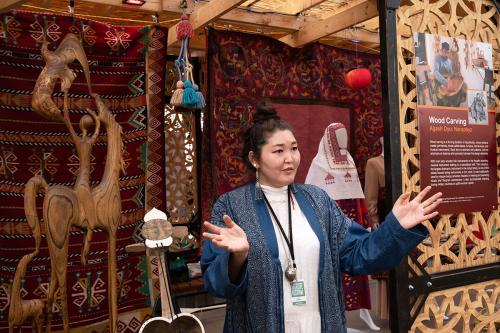 A woman gestures with both hands outward in front of a display of woven textiles, carved wooden figures, wooden instruments, and clothing on mannequins.