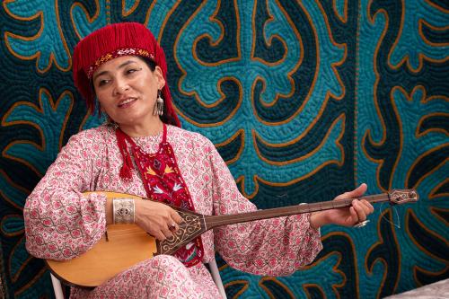 A woman plays a two-string wooden lute, sitting in front of a turquoise quilt with yellow embroidery.