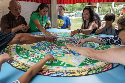 Several people, standing around the circular mandala on a table under a tent, reach their hands toward the center of the mandala.