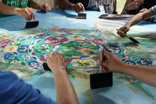 In a circle, several hands use foam brushes to brush the sand of the mandala, now blurred around the edges, toward its center.