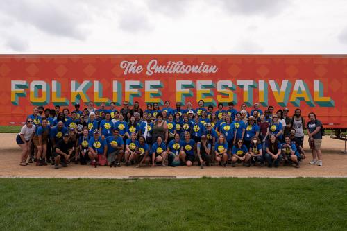 A large group of people in blue T-shirts stand in front of a large trailer, smiling at the camera. The trailer is painted with an orange background and light orange diamonds and squares. The text on the trailer reads “The Smithsonian Folklife Festival.”