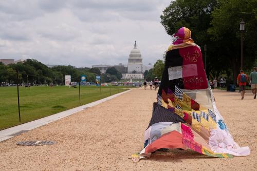 Wearing a flowing, quilted cape, a person walks down a gravel path toward the U.S. Capitol Building, gray clouds overhead.
