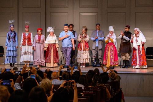 Ten models pose on stage, wearing elaborates dresses, vests, shirts, and headdresses, in front of a seated audience. Another man in front of them prepares to take a bow.