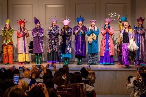 On stage, people in elaborate outfits in purple, teal, pink, and orange line up and applaud, while a woman in front tosses a handful of candy toward the seated audience.