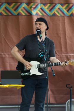 A man wearing all black plays a black electric guitar on stage.