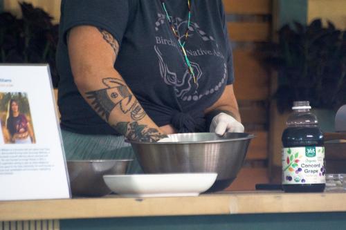 Close-up on a person with tattooed arms, one latex-gloved hand in a silver mixing bowl. To the side, a bottle of Concord grape juice. 