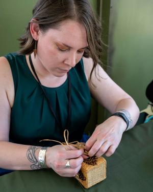 A woman with chin-length brown hair, green tank top, and silver jewelry works at a table hand-weaving a box-shaped  basket.