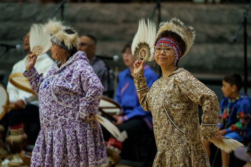 Two women wearing patterned regalia dancing while holding feathered fans.