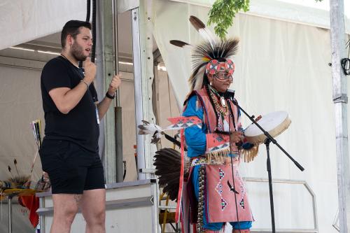 An ASL interpreter stands alongside a man in Native regalia and tall feathered headdress, holding a handdrum, signing.