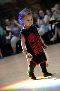 A young child wearing a black smock with red trim and design walks in a pool of rainbow light on stage.