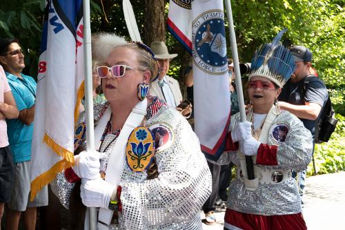A woman wearing silver sequined regalia carries a flag.