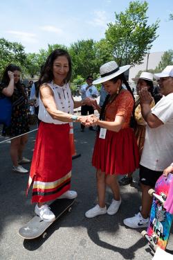 A woman in a red dress holds the hands of another woman wearing a white dress who is riding a skateboard.