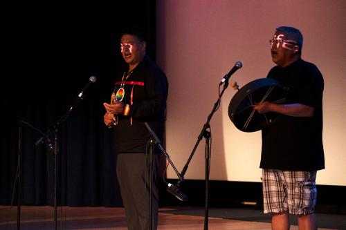 Two men on a dimly lit stage, singing and playing a wooden rattle and a frame drum.