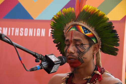 The man in the feathered crown, now with black lines painted on his cheeks and a red line down the center of his nose, and his chin painted red, sings into the microphone.