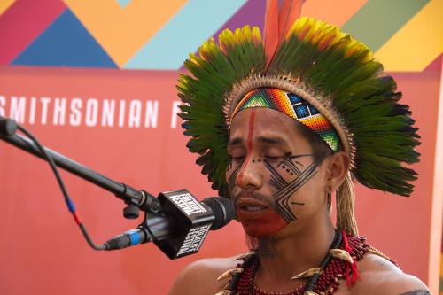 A man with black and red face paint wearing a headdress made of leaves speaks into a microphone.