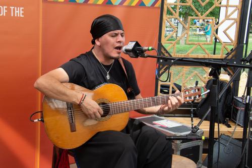 A man wearing all  black clothes and a black headwrap sits on an outdoor stage, singing and playing acoustic guitar.