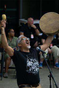 A man with gray hair and a black shirt shouting and holding a drum and drumstick above his head.