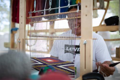 Through the warp of a loom, a man in a white Levi’s shirt smiles and he weaves a geometric pattern in purple, red, white, and gold.