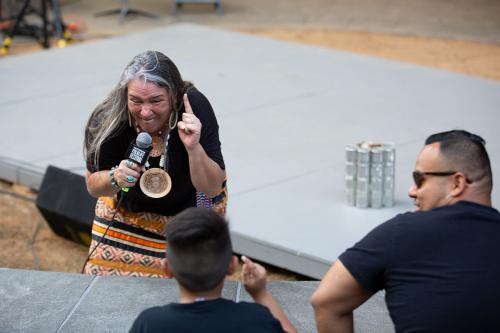 A woman with long, gray hair crouches down to face a young boy. They both raise their pointer fingers. A man next to the boy looks over at him, smiling.