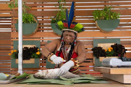 The woman in the feathered crown and beaded jewelry works in an outdoor kitchen, squeezing lemon into a gutted whole fish.