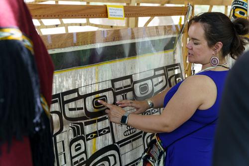 A woman points at a black design printed on a clear plastic sheet, laid over the warp of weaving in progress.