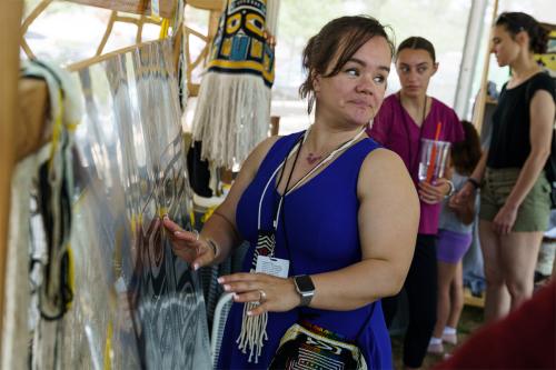 A woman in a blue dress and beaded jewelry looks over her shoulder while touching a plastic overlay with a black design stretched over a loom.