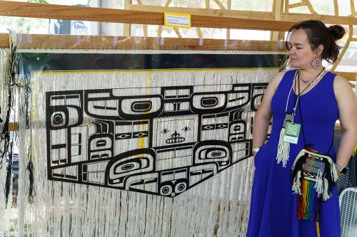 A woman poses next to her weaving in progress.