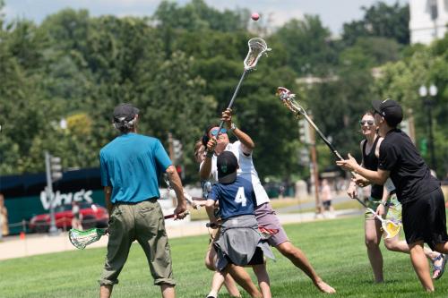 Six people, adults and children, running in the grass and wielding lacrosse sticks. One person is about to catch a small red ball in mid-air.