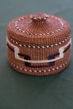 Round woven basket with lid in a reddish hue, with white and dark brown pattern detail.