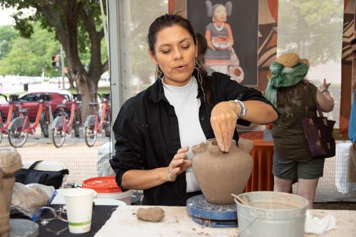A woman forms one of four spouts on a light brown clay jug shape.