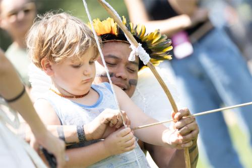 A man wearing a feather headdress guides a young boy in holding a bow and arrow.