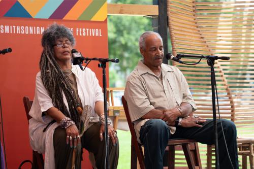 A woman with long braids wearing a white cardigan and a man wearing a tan shirt sit on a stage, eyes closed in prayer.