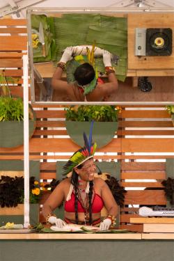 A woman wearing a red top and colorful headdress prepares a whole fish in an outdoor demonstration kitchen.