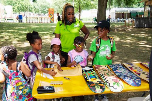 Woman in a green polo shirt and five children playing with miniature skateboards and looking at a display of skateboards with painted decks.